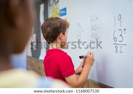 Rear view of young boy solving addition and subtraction on white board at school. Schoolboy thinking while solving math's sum. Child writing the solution of the mathematical operation in classroom.