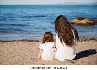 Rear View Of A Young Brunette Mom Sitting Next To Her Daughter While Looking At The Ocean