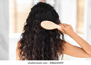 Rear View Of Young Brunette Woman Combing Her Long Curly Hair With Bamboo Brush While Standing In Bathroom, Back Shot Of Unrecognizable Female Making Morning Beauty Routine At Home, Copy Space