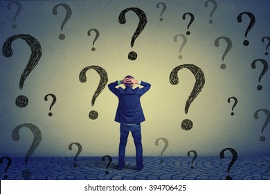 Rear view of young business man with hands on head standing in front of wall with many questions wondering what to do next. Full length of businessman facing the wall. Job work challenge concept 