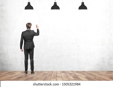 Rear view of young businessman in dark suit writing with pen on concrete mock up wall. Concept of business planning and education