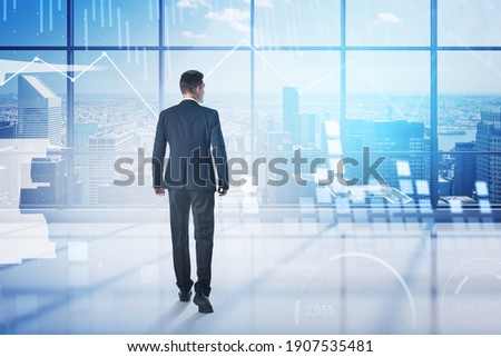 Rear view of young businessman walking in his panoramic office with double exposure of blurry financial graphs. Toned image