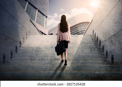 Rear view of young businesswoman carrying a suitcase while walking upward on the stairs with skyscrapers background - Powered by Shutterstock