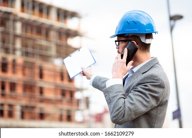 Rear View Of A Young Caucasian Architect Or Engineer Wearing Casual Business Clothes Inspecting Construction Site And Talking On The Phone