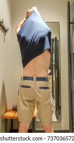 Rear View Of A Young Caucasian Handsome Man Taking Off T-shirt In Front Of A Mirror In A Changing Room Of A Clothing Store