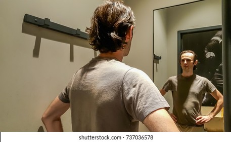 Rear View Of A Young Caucasian Handsome Man Standing In Front Of A Mirror In A Changing Room Of A Clothing Store