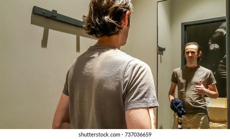 Rear View Of A Young Caucasian Handsome Man Standing In Front Of A Mirror In A Changing Room Of A Clothing Store