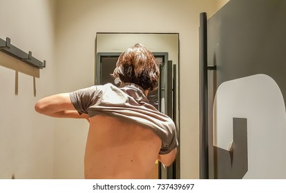 Rear View Of A Young Caucasian Handsome Man Taking Off T-shirt In Front Of A Mirror In A Changing Room Of A Clothing Store