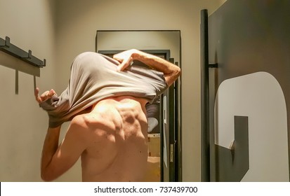 Rear View Of A Young Caucasian Handsome Man Taking Off T-shirt In Front Of A Mirror In A Changing Room Of A Clothing Store