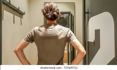 Rear View Of A Young Caucasian Handsome Man Standing In Front Of A Mirror In A Changing Room Of A Clothing Store