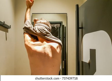 Rear View Of A Young Caucasian Handsome Man Taking Off T-shirt In Front Of A Mirror In A Changing Room Of A Clothing Store