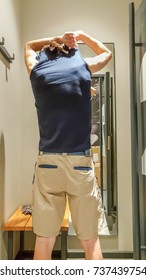 Rear View Of A Young Caucasian Handsome Man Taking Off T-shirt In Front Of A Mirror In A Changing Room Of A Clothing Store