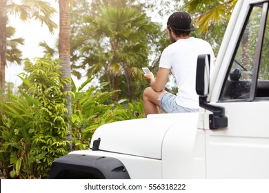 Rear view of young Caucasian man sitting atop of his white four-wheel drive vehicle, taking picture of beautiful wild nature around him using mobile phone while having vacation in exotic country - Powered by Shutterstock