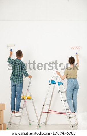 Rear view of young couple in casual clothing standing on stepladders and paining wall with paint roller while making interior of dream in new flat
