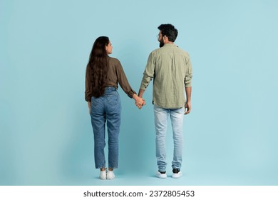 Rear view of young eastern couple bearded guy and long-haired woman walking over plain blue background, holding hands, looking at each other and smiling, full length. Love, affection, relationships - Powered by Shutterstock