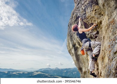 Rear view of young female climber climbing with rope and carbines, looking for next grip on a big rocky wall against blue sky and mountains. Summer time. Climbing equipment