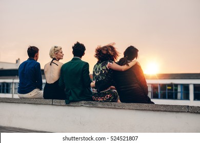Rear view of young friends relaxing together on rooftop at sunset. Young men and women sitting on terrace in evening.