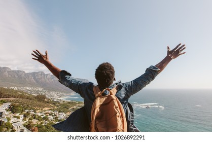 Rear view of young guy with backpack standing outdoors with arms spread open against seascape. Man enjoying the view from the top of the mountain.