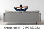 Rear view of young guy sitting on comfortable couch at home in living room, looking at wall. Casual man relaxing on sofa, leaning back holding hands behind head, enjoying weekend free time or break