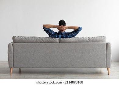 Vista trasera de un joven sentado en un cómodo sofá de casa en la sala de estar, mirando a la pared. El hombre informal se relaja en el sofá, apoyándose en la parte trasera, disfrutando del tiempo libre del fin de semana o descansando