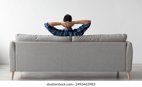 Rear view of young guy sitting on comfortable couch at home in living room, looking at wall. Casual man relaxing on sofa, leaning back holding hands behind head, enjoying weekend free time or break - Powered by Shutterstock