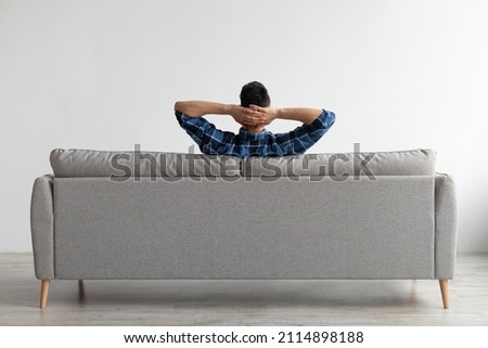 Rear view of young guy sitting on comfortable couch at home in living room, looking at wall. Casual man relaxing on sofa, leaning back holding hands behind head, enjoying weekend free time or break