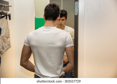 Rear View Of A Young Handsome Man Trying On Clothes In Clothing Store's Changing Room In Front Of A Mirror Or In Room Closet