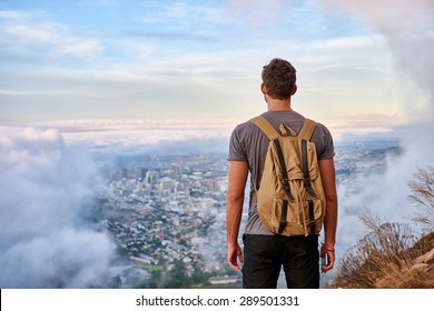 Rear View Of A Young Hiker Standing On A Mountain Path Looking Out Over The City Through The Clouds