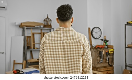 Rear view of a young hispanic man in a checkered shirt standing thoughtfully in an organized carpentry workshop. - Powered by Shutterstock