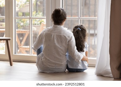 Rear view young loving mother hugging little daughter sitting on warm floor near window looking together outside, talking, admire view from modern house, enjoy leisure at cozy home. Tenancy concept - Powered by Shutterstock