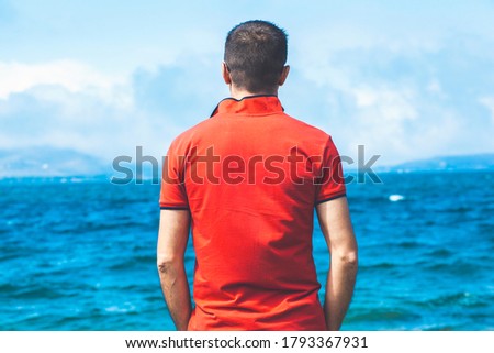 Rear view of young male traveler in shirt and shorts standing by the sea and enjoying nature and seclusion on a summer day
