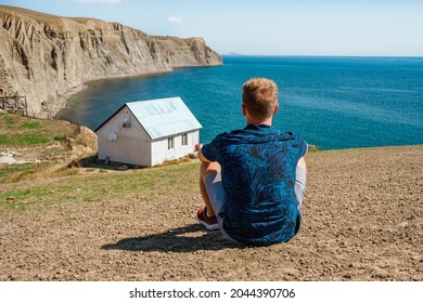 Rear view a young man in front of a white lonely house on the edge of a cliff with a picturesque mountain landscape and a view of the sea