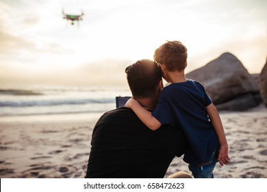 Rear view of young man and little boy operating the drone by remote control at the beach. Father and son flying drone at the sea shore, enjoying summer vacation. - Powered by Shutterstock