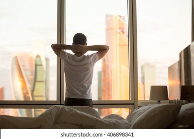 Rear view of young man looking at dawn city scenery in window after waking up. Handsome casual guy relaxing in penthouse in the morning with hands crossed behind his head. Motivation concept - Powered by Shutterstock