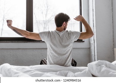 Rear View Of Young Man In Pajamas Stretching While Sitting In Bed In The Morning