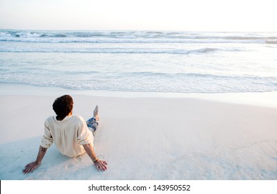 Rear view of a young man sitting on a white sand beach during sunset contemplating the scenery and the blue sea waves during his vacation in an idyllic nature scene destination. - Powered by Shutterstock