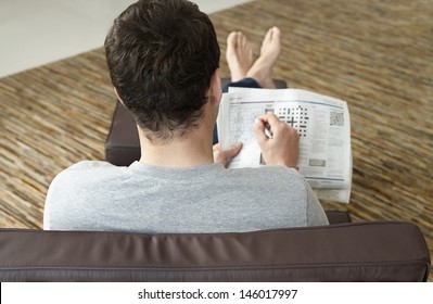 Rear View Of A Young Man Sitting On Sofa And Doing Crossword Puzzle In Newspaper