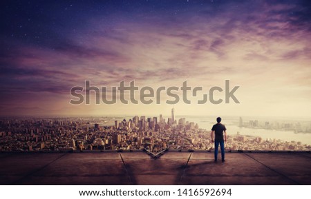 Rear view of young man standing on the rooftop of a skyscraper watching sunset over the big city horizon. Businessman on the roof getting inspired and motivated.
