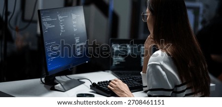 Rear view of a young programmer intensely reviewing code displayed on a monitor in a dark office. Software engineer typing and debugging on program. App development, hacker culture, and cybersecurity