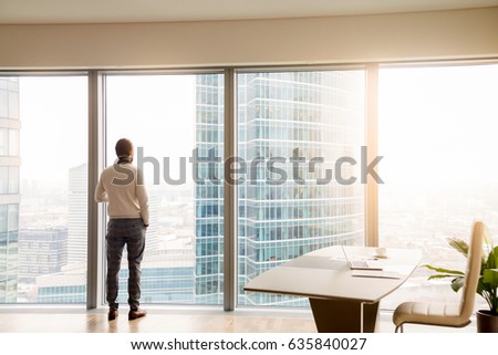 Rear view at young successful businessman standing in office looking through full-length window at cityscape with skyscrapers, dreaming or resting, waiting for meeting or considering business offer
