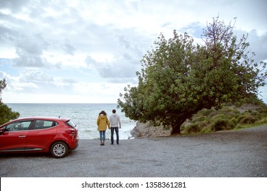 Rear view of young tourist couple standing on rocks near his car on a winter holiday with coats, looking at the sea. Travel lifestyle exterior. - Powered by Shutterstock