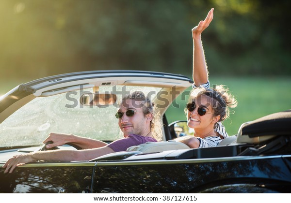 Rear view. Young\
trendy couple happy to drive his convertible car on a country road\
in summer. Shot with flare