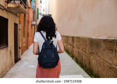 Rear View Of A Young Woman With A Backpack Walking Down A Lonely City Alley Alone. Sense Of Danger