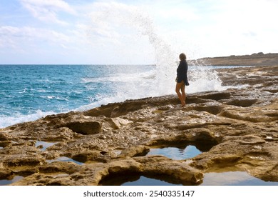 Rear view of a young woman in black swimsuit and black shirt on the standing on the rocky sea shore. Big wave splash in the background. - Powered by Shutterstock