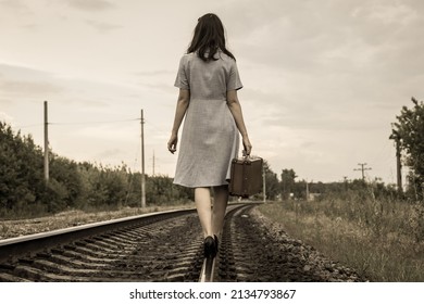 A rear view of a young woman in a dress and with a suitcase walking away along the rails of a railway road. The concept of departure, emigration, departure from the country, refugee status,deportation - Shutterstock ID 2134793867
