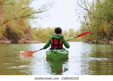 Rear view of young woman in green kayak paddle at river near trees with gentle green leaves at spring