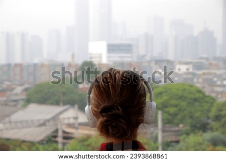 Rear view of a young woman with headphones on her ears looking at a blurry city view.