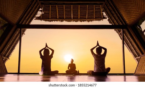 Rear view of young woman with her daughter practicing yoga while sitting on mattress near a Buddha statue with dusk sky view background on the window hotel - Powered by Shutterstock