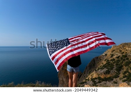 Rear view of a young woman holding an American flag waving at the coastline against the sunny bright sea.