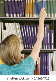 Rear View Of A Young Woman Reaching For Book From Library Shelf